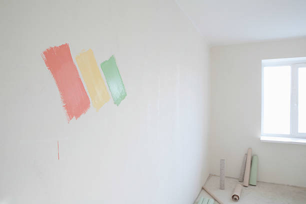 Внутренняя стена комнаты с тремя образцами краски разного цвета, нанесенными рядом с окном, на полу видны принадлежности для рисования.